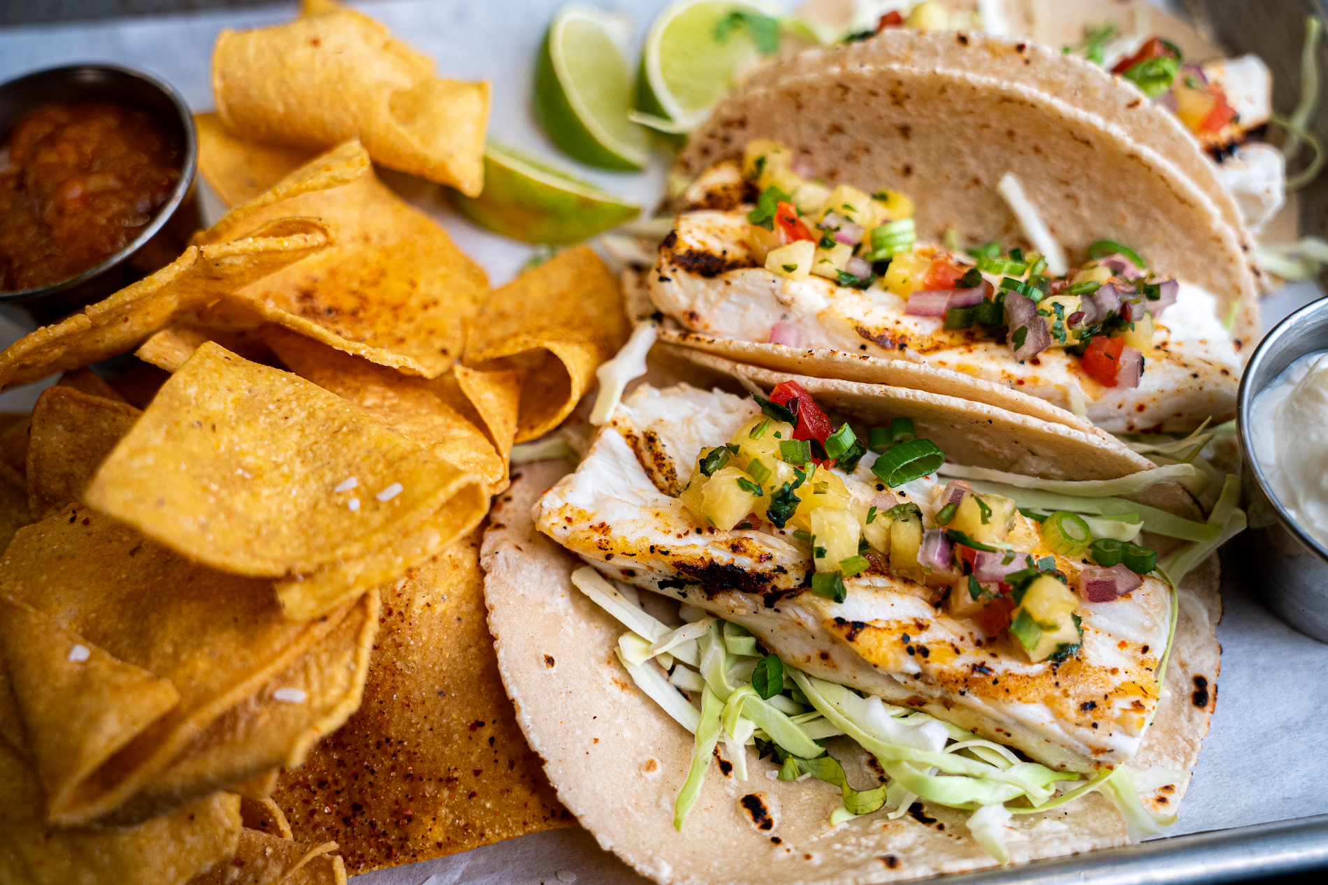 Two grilled fish tacos with cabbage, pineapple salsa, and scallions on tortillas, served with tortilla chips, lime wedges, salsa, and a side of sauce on a metal tray.