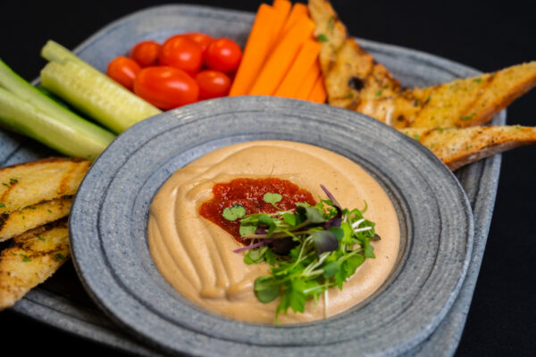 A plate with hummus topped with red sauce and greens, surrounded by cucumber sticks, cherry tomatoes, carrot sticks, and slices of toasted bread on a blue-gray dish.
