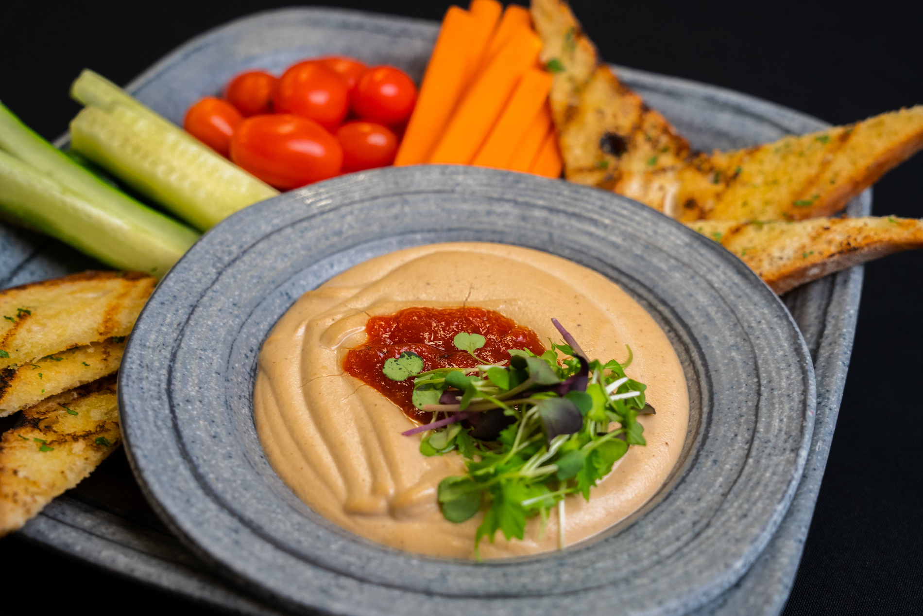 A plate with hummus topped with red sauce and greens, surrounded by cucumber sticks, cherry tomatoes, carrot sticks, and slices of toasted bread on a blue-gray dish.