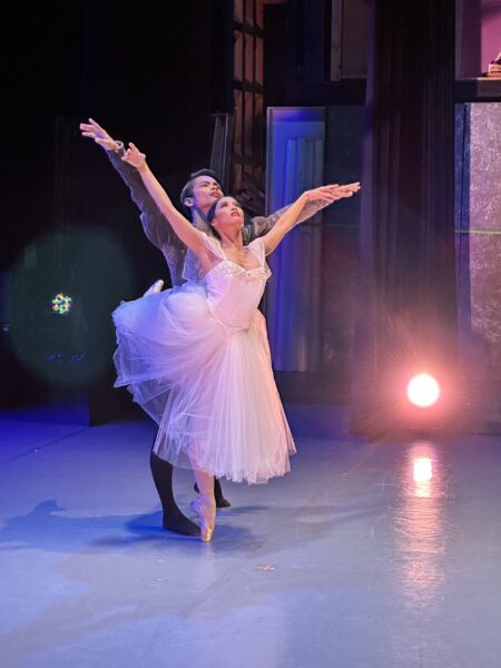 Two ballet dancers perform on stage. The female dancer is en pointe in a white tutu, arms raised gracefully. The male dancer stands behind, supporting her, both looking upward. Stage lights and a dramatic backdrop create a theatrical scene.
