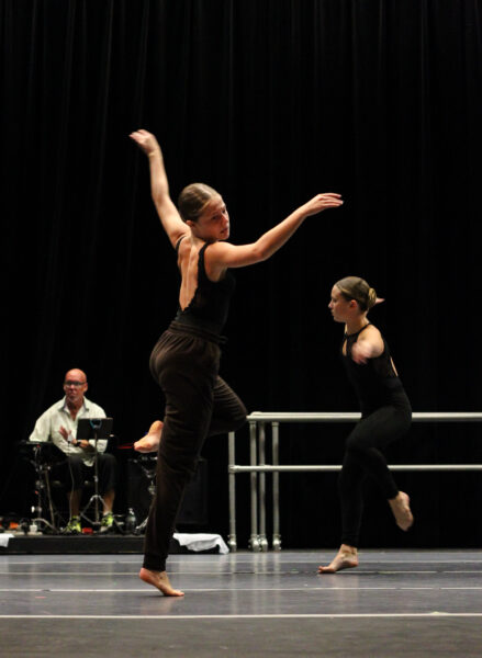Two women in black dance barefoot on stage, striking expressive poses. A man in white sits in the background playing an electronic instrument against a dark backdrop.