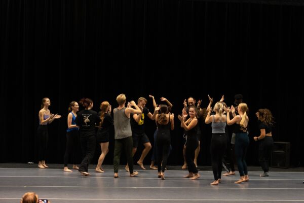 A group of people in dance attire stand in a circle on stage, clapping and raising their arms, with a dark curtain as the background.