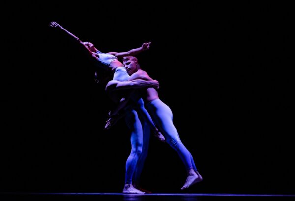 Two dancers in matching light blue bodysuits perform a lift on stage, with one dancer holding the other aloft against a dark background, creating a dramatic and graceful pose.