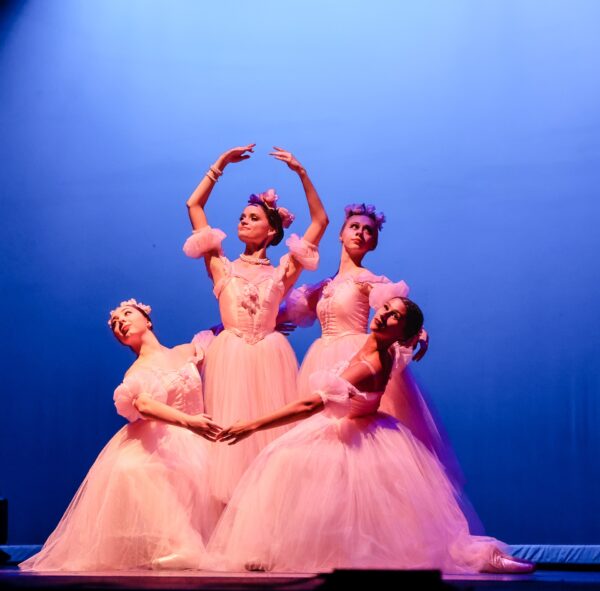 Four ballerinas in pink tutus pose gracefully on stage against a blue backdrop, with three kneeling and one standing with arms raised. All wear flower headpieces and delicate expressions.