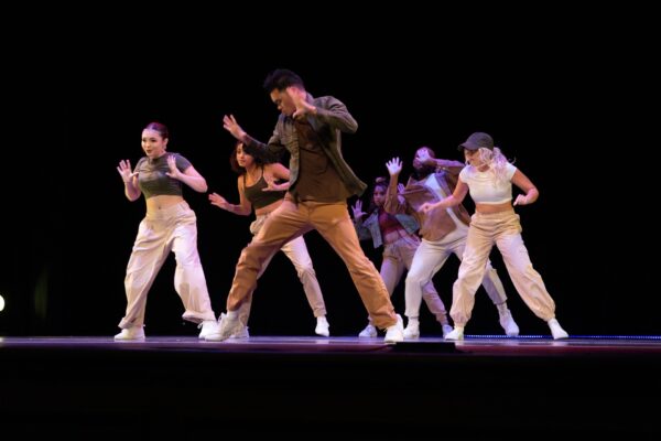A group of six dancers perform a hip hop routine on stage, all dressed in casual, coordinated outfits, moving energetically in sync under stage lights against a dark background.