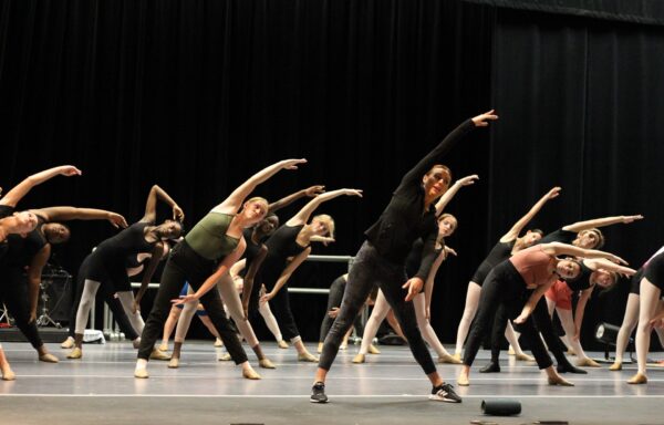 A group of ballet dancers in a studio stand in a row, stretching to one side with one arm raised overhead. Most are wearing leotards and tights, following the instructor at the front. The background is dark with curtains.