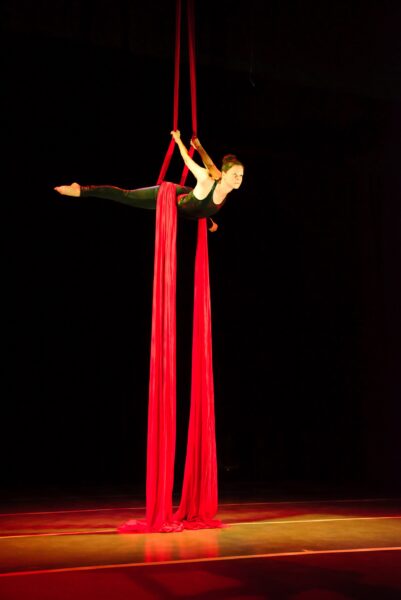 A performer in a black outfit balances horizontally in the air, holding onto long red aerial silks during an acrobatic routine on a dimly lit stage.