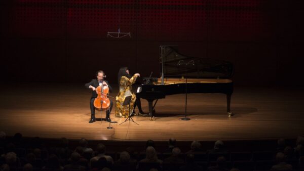 A cellist and a pianist perform on stage under warm lighting in a concert hall, with an audience seated in the foreground. The pianist wears a gold outfit, and the cellist plays beside a grand piano.