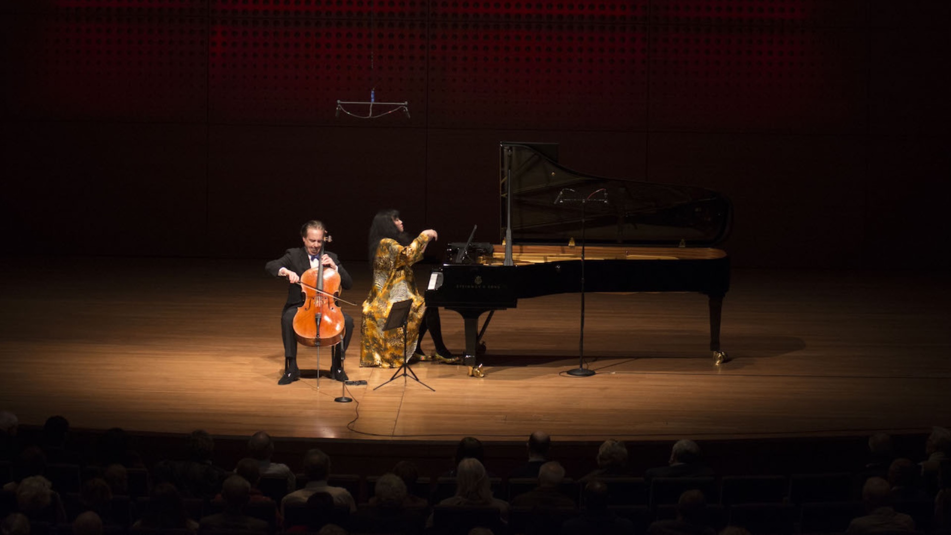A cellist and a pianist perform on stage under warm lighting in a concert hall, with an audience seated in the foreground. The pianist wears a gold outfit, and the cellist plays beside a grand piano.