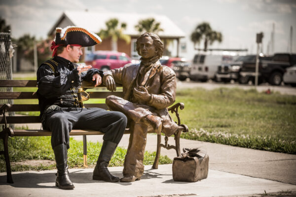 A man in a pirate costume sits on a bench, gesturing as if talking to a bronze statue of a seated person. A small bronze bag rests on the ground beside the bench, with cars and palm trees in the background.