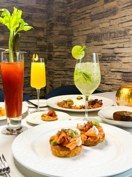 A table set with various brunch dishes, including smoked salmon bruschetta, a tomato cocktail with celery and pickle, a mimosa, a lime cocktail, and assorted plates against a stone wall background.