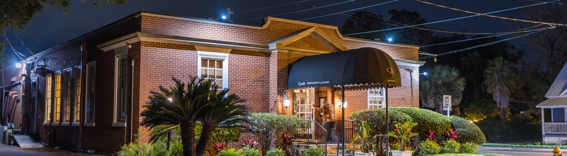 A small brick building with a black awning, outdoor lights, green shrubs, and palm plants, seen at night on a quiet, empty street.