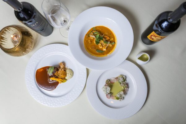 Three elegantly plated gourmet dishes are arranged on a white tablecloth, accompanied by two bottles of red wine, a glass of white wine, a lit candle, and a small dish of olive oil.