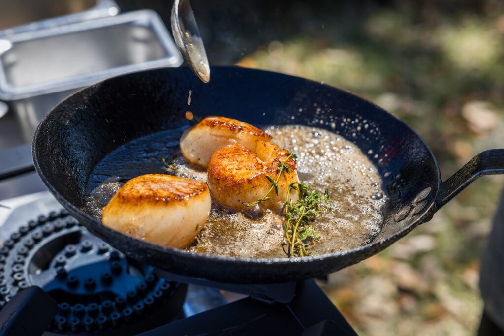 Three seared scallops cook in a black pan with bubbling butter and fresh herbs while someone drizzles more butter onto them with a spoon.