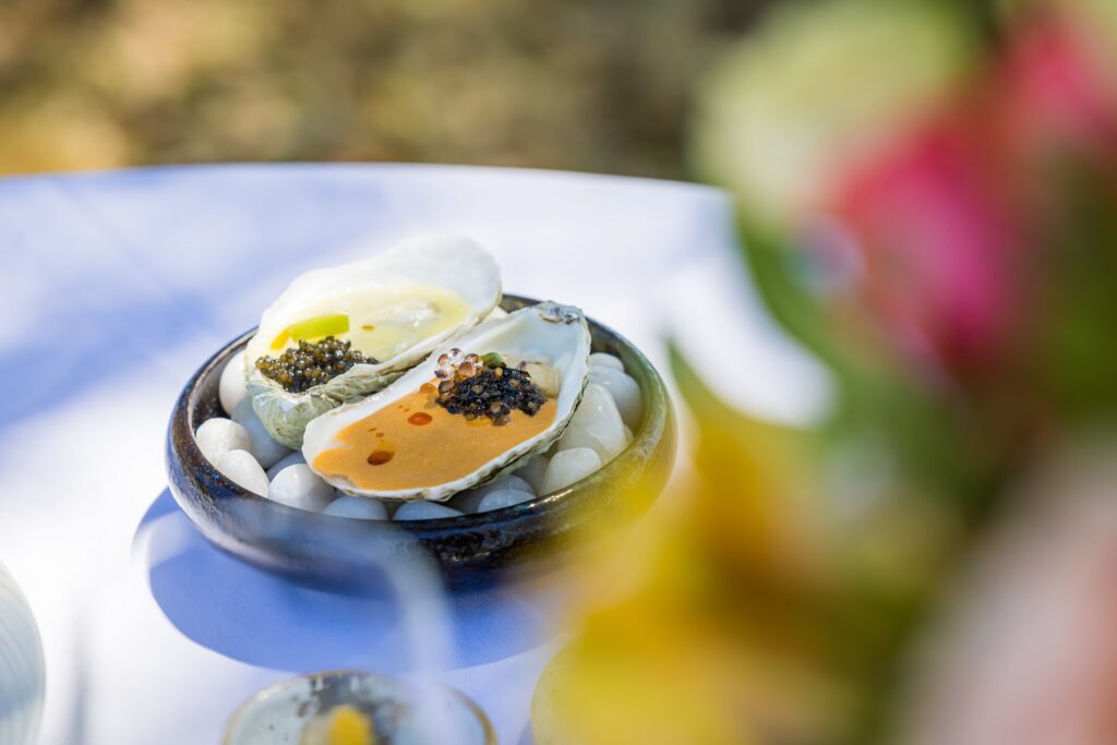 Two half-shell oysters topped with caviar and garnishes sit on white stones in a black bowl, placed on a white tablecloth. The background is blurred with colorful flowers visible on the right side.