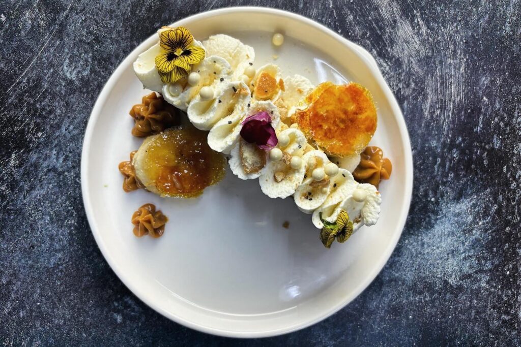 A white plate with a gourmet dessert topped with whipped cream, caramelized rounds, edible flowers, and caramel dollops, set on a dark textured surface.