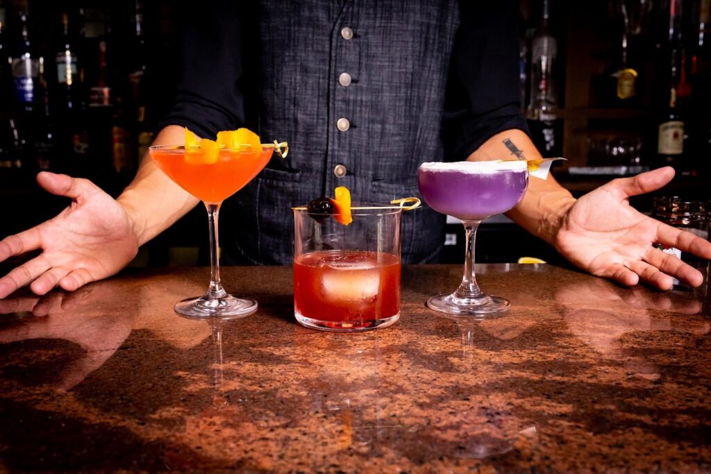 A bartender stands behind a bar with three colorful cocktails: an orange drink, a red drink with a cherry, and a purple drink with a lemon twist, arms open in a welcoming gesture.
