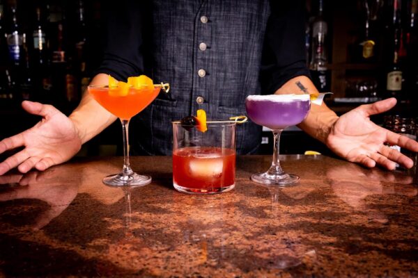 A bartender stands behind a bar with three colorful cocktails: an orange drink, a red drink with a cherry, and a purple drink with a lemon twist, arms open in a welcoming gesture.