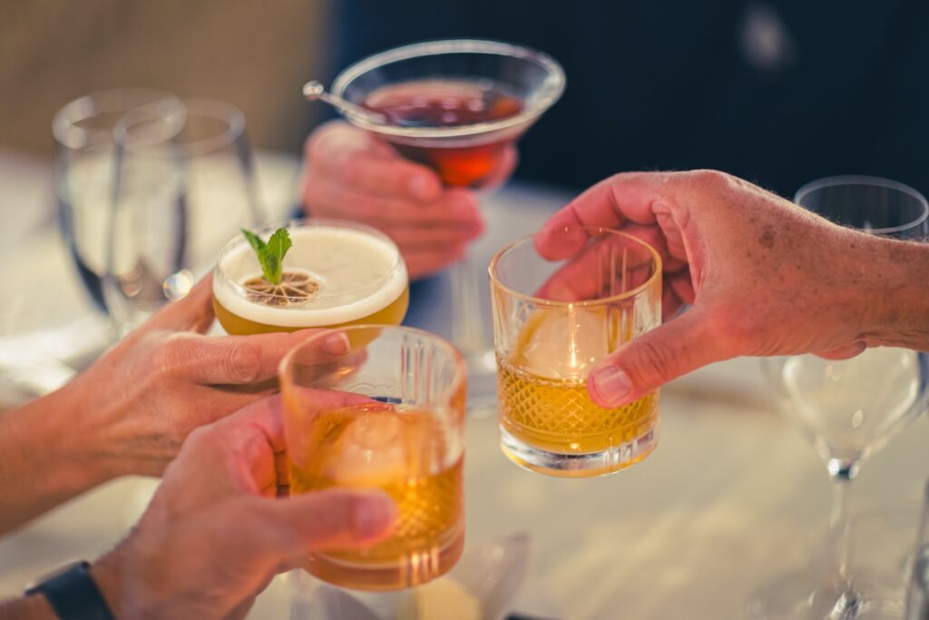 Four people clinking different cocktails and drinks together over a table, celebrating or toasting, with glasses and blurred background visible.