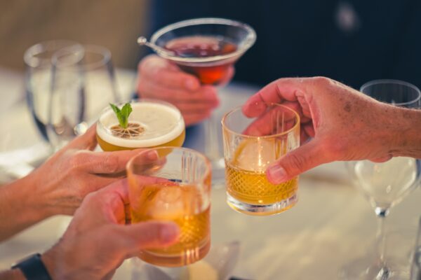 Four people clinking different cocktails and drinks together over a table, celebrating or toasting, with glasses and blurred background visible.