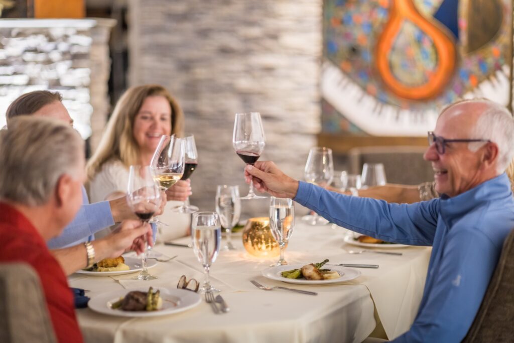 Four people sit at a restaurant table, smiling and raising glasses in a toast. Plates of food and glasses of wine and water are on the white tablecloth, with a candle and colorful wall art in the background.