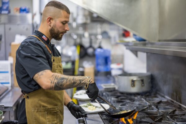 A chef wearing black gloves and a brown apron cooks food in a frying pan over a gas stove in a commercial kitchen, plating a dish with focused attention.