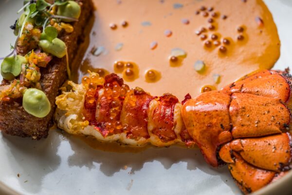 Close-up of a lobster tail with orange sauce, garnished with what appears to be salmon roe and microgreens, served beside a crispy, toast-like item topped with diced vegetables and a green creamy sauce.