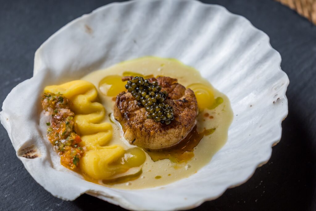 A seared scallop topped with black caviar sits on a shell, accompanied by a yellow purée, a colorful vegetable relish, and a light sauce.