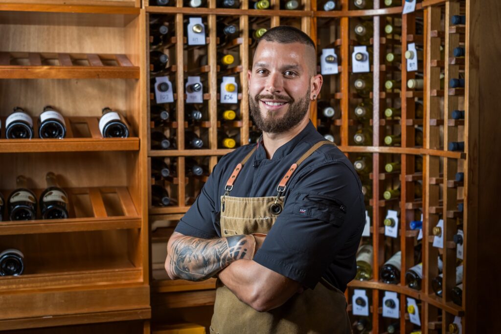 A man in a chefs uniform with tattooed arms stands smiling and arms crossed in front of shelves filled with wine bottles in a wooden wine cellar.