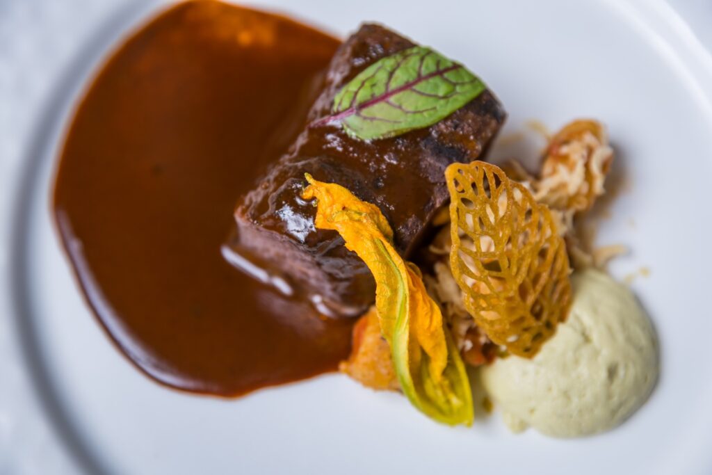 A gourmet dish features a glazed meat portion with brown sauce, a green-veined leaf, yellow edible flower, crispy garnish, and a dollop of creamy puree on a white plate.