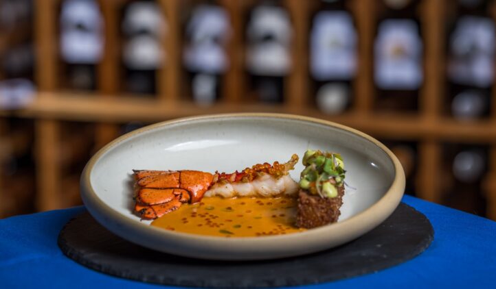 A gourmet seafood dish featuring lobster tail, a delicate sauce, and a small garnished cube, artfully presented on a white plate with a blurred wine rack in the background.