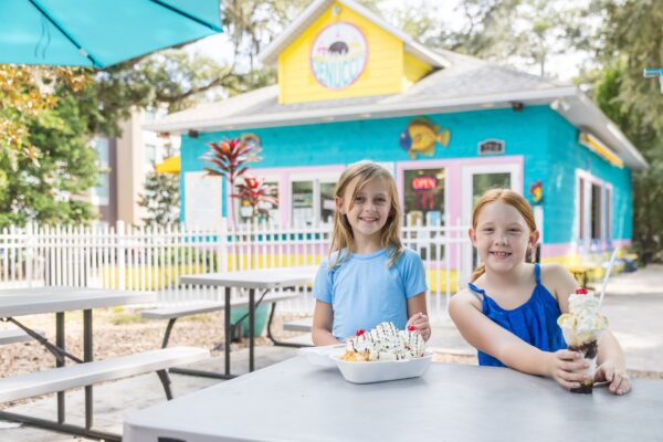 Two smiling young girls sit at an outdoor table with ice cream treats in front of a colorful, blue and yellow ice cream shop on a sunny day.