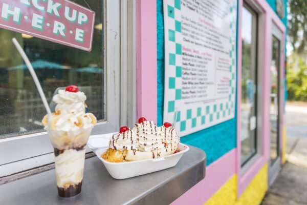 A banana split sundae and a tall ice cream parfait with whipped cream and cherries are placed on a counter outside a colorful ice cream shop, with a menu and Pick Up Here sign visible.