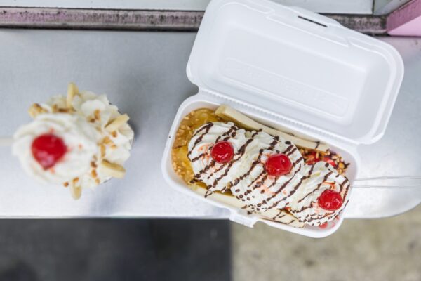 A banana split in a white takeout container topped with whipped cream, cherries, chocolate sauce, and sprinkles sits on a counter next to a cup of soft serve ice cream with whipped cream and a cherry.