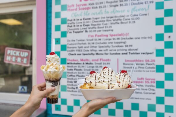Two hands hold desserts in front of an ice cream shop menu: one holds a tall parfait with whipped cream and a cherry, the other a large banana split topped with whipped cream, chocolate syrup, and cherries.
