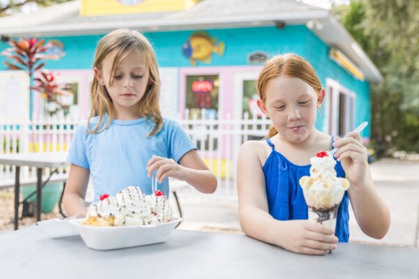 Two young girls sit at an outdoor table enjoying ice cream treats; one has a large sundae with whipped cream and cherries, and the other has a parfait with whipped cream, in front of a colorful ice cream shop.