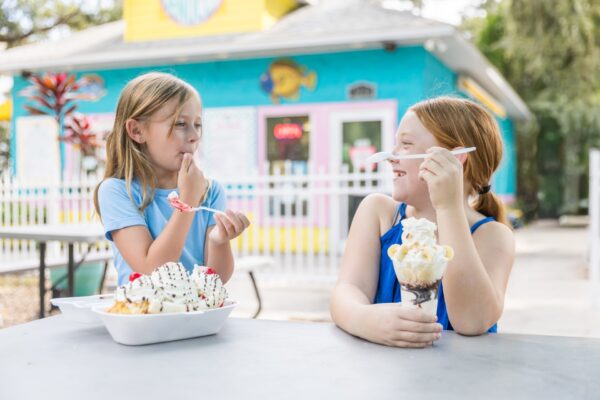 Two young girls sit at an outdoor table enjoying ice cream treats, smiling and laughing together, with a colorful ice cream shop in the background on a sunny day.