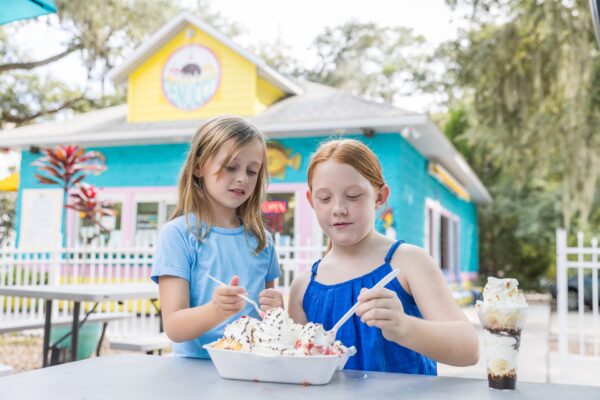 Two young girls sit at an outdoor table sharing a large ice cream sundae with whipped cream and cherries. An ice cream shop with a bright yellow and blue exterior is in the background. A tall ice cream parfait is on the table.
