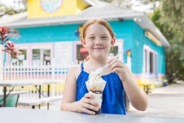 A young girl with red hair wearing a blue dress smiles while holding a sundae topped with whipped cream, sitting at an outdoor table in front of a colorful ice cream shop.