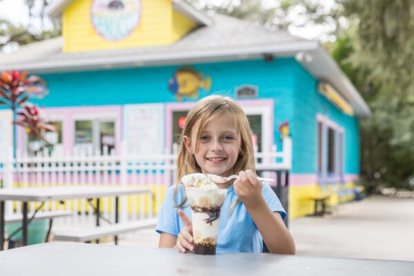 A smiling young girl sits at an outdoor table in front of a colorful ice cream shop, holding a large ice cream sundae and scooping it with a spoon.