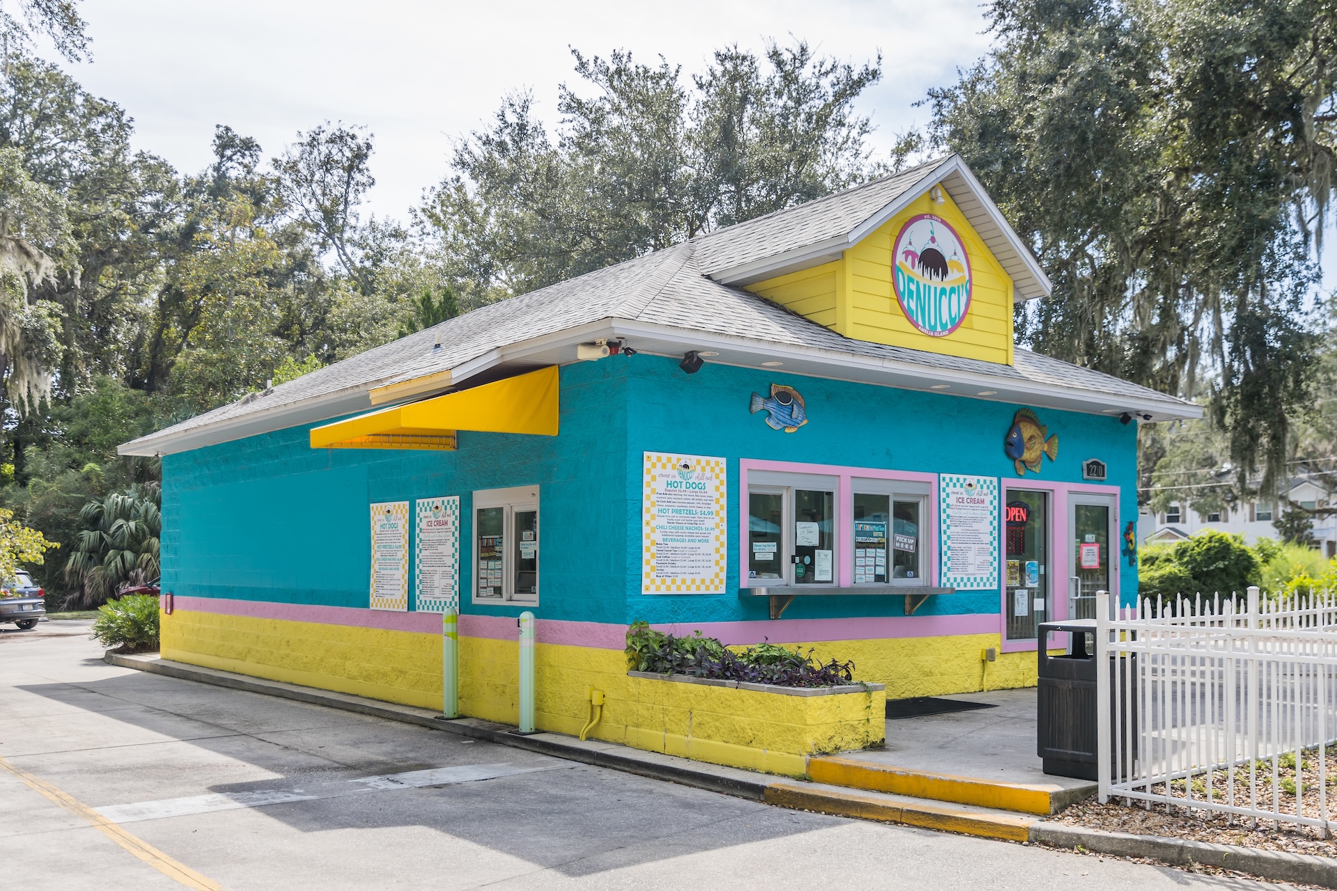 A small, colorful restaurant with teal, yellow, and pink walls, bright signage, and several menu boards displayed outside. The building has a tropical theme and is surrounded by trees and a paved area.
