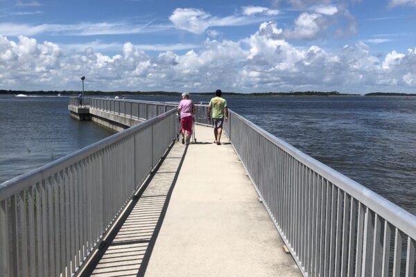 Two people walk side by side on a concrete pier with metal railings, surrounded by water under a partly cloudy sky. A dock extends to the left, and land is visible in the distance.