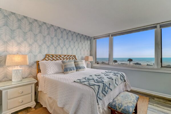 A cozy bedroom with a large bed, patterned wallpaper, two bedside tables with lamps, and a window offering a stunning ocean view under a clear blue sky.