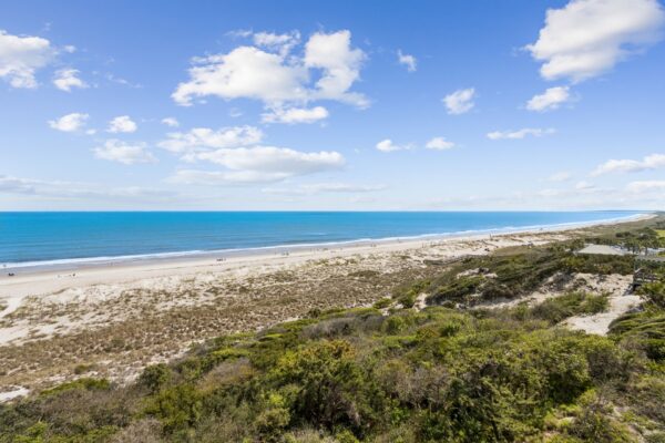 A wide sandy beach bordered by grassy dunes and green shrubs, with turquoise ocean waves and a bright blue sky filled with scattered white clouds.