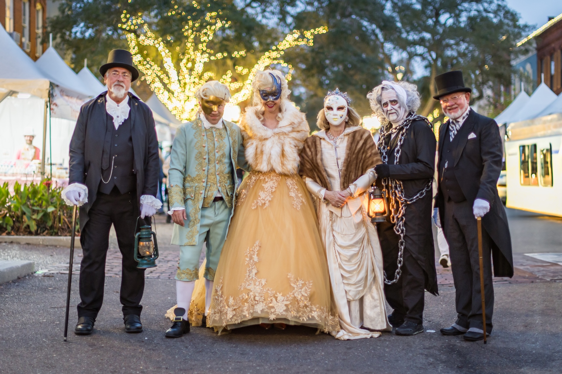 Six people in elaborate historical and masquerade costumes pose outdoors at dusk, with festive lights and tents in the background. Some hold lanterns, and two wear white face makeup.