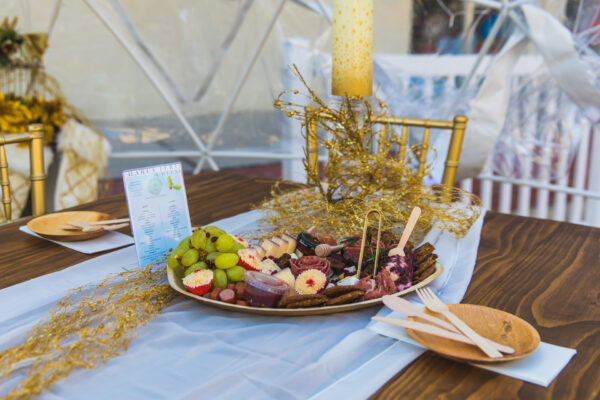 A wooden table set for a meal features a decorative centerpiece, a menu, and a platter with assorted cheeses, grapes, meats, and crackers. Wooden plates, cutlery, and a gold chair are also visible.