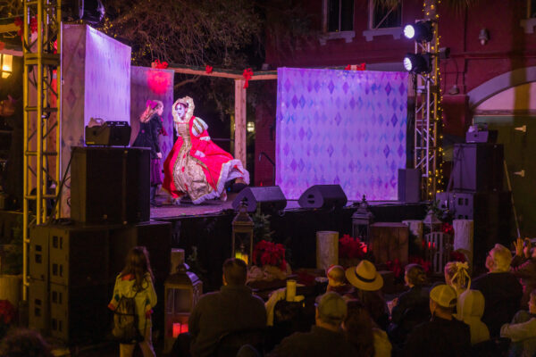 A performer in an elaborate red and white Renaissance-style costume dances on an outdoor stage at night, while an audience watches. The stage is decorated with string lights and festive holiday decor.