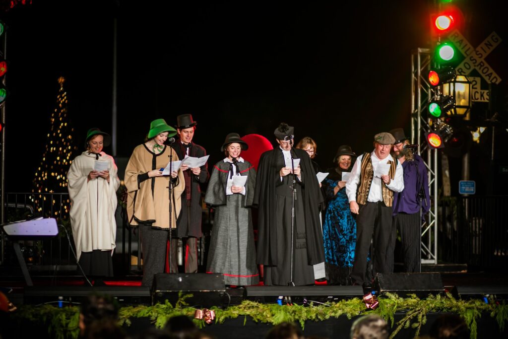 A group of people dressed in Victorian-era costumes stand on a stage singing or reading from papers, with festive lights and a decorated Christmas tree in the background.