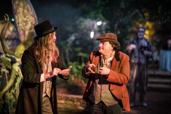 Two people in old-fashioned costumes gesture animatedly outdoors at night, with a blurred figure in costume standing in the background near trees and a statue.