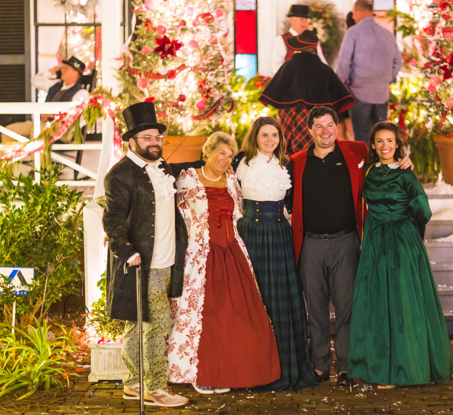 Five people dressed in festive, historical costumes smile and pose together outdoors at night, surrounded by holiday decorations and lights. Other people and plants are visible in the background.